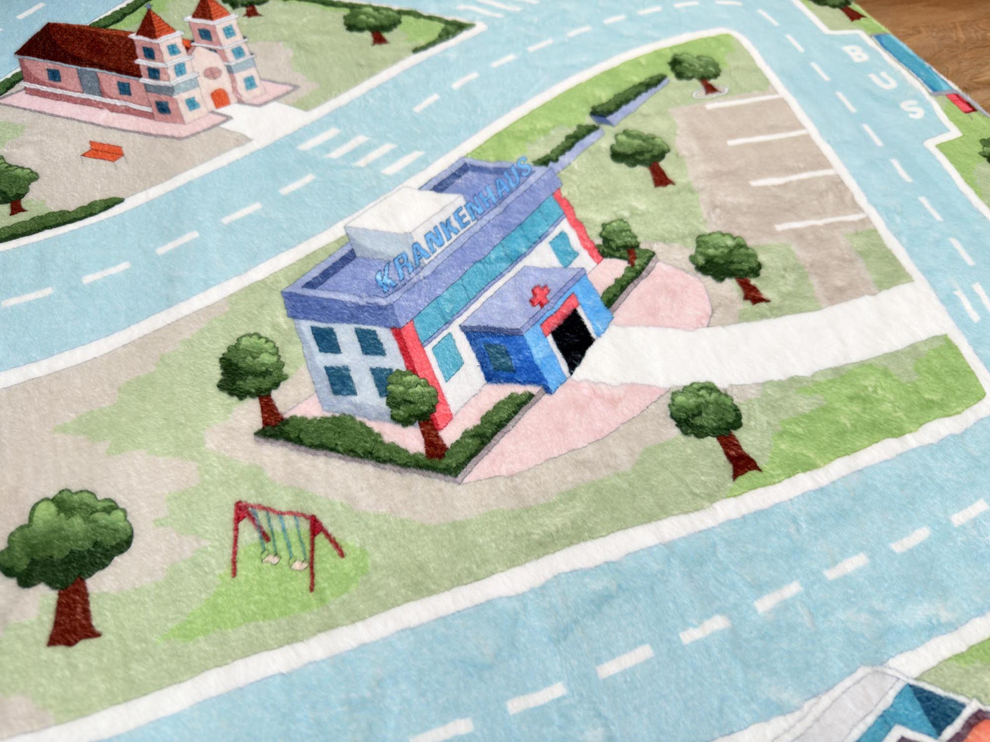 Fluffy Town Spielteppich – Spielbereich mit Krankenhaus und Schaukeln Krankenhaus und Spielplatz auf dem Fluffy Town Spielteppich mit umliegender Straße