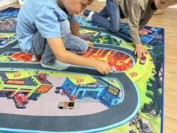 Zwei Kinder spielen mit Autos auf dem Spielteppich „Monster City XL“ 200×140 cm mit Lavafeld, Werkstatt und Feuerwehr